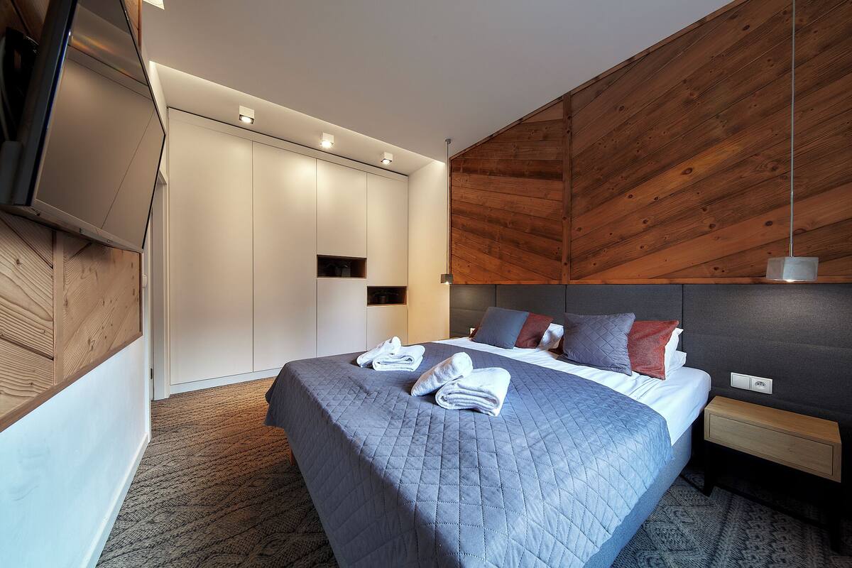A modern hotel room with a large bed, a TV, a wardrobe, and a wooden accent wall.