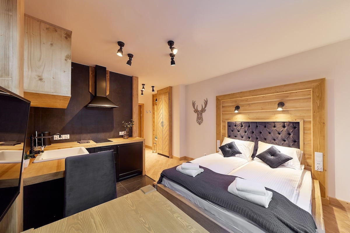 A modern hotel room with a wooden headboard, a large bed with pillows, a desk with a chair, and a kitchenette.
