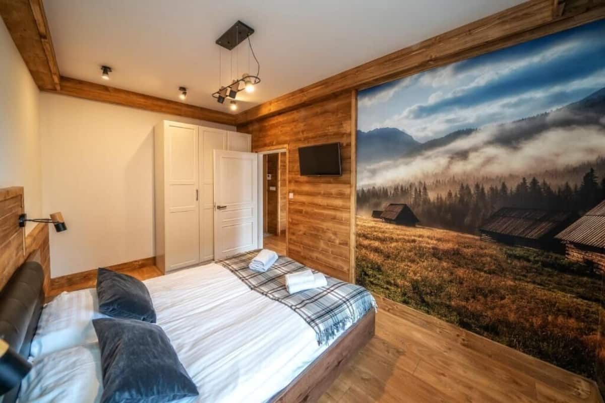 A bedroom with a bed, a white wardrobe, a mounted TV, and a large window with a forest and mountain view.