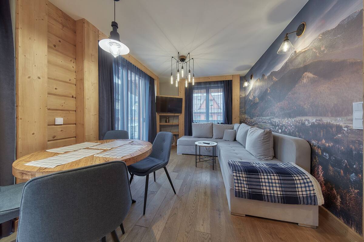 A cozy living room with a wooden interior, a round dining table with chairs, a sofa with a plaid throw, and a mural of a mountainous landscape on the wall.