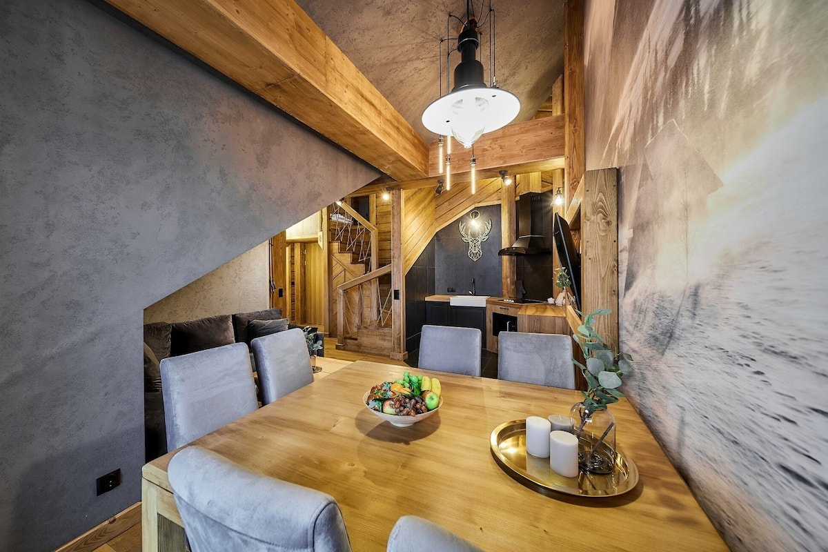 A dining area with a wooden table, grey upholstered chairs, a fruit bowl, and a decorative plant.
