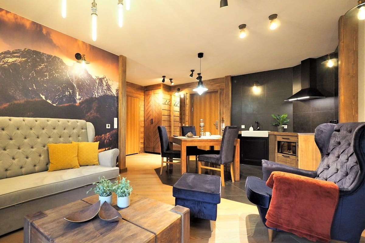 A modern living room with a sofa, coffee table, and chairs, a dining area with a table and chairs, and a kitchen with a stove and sink.
