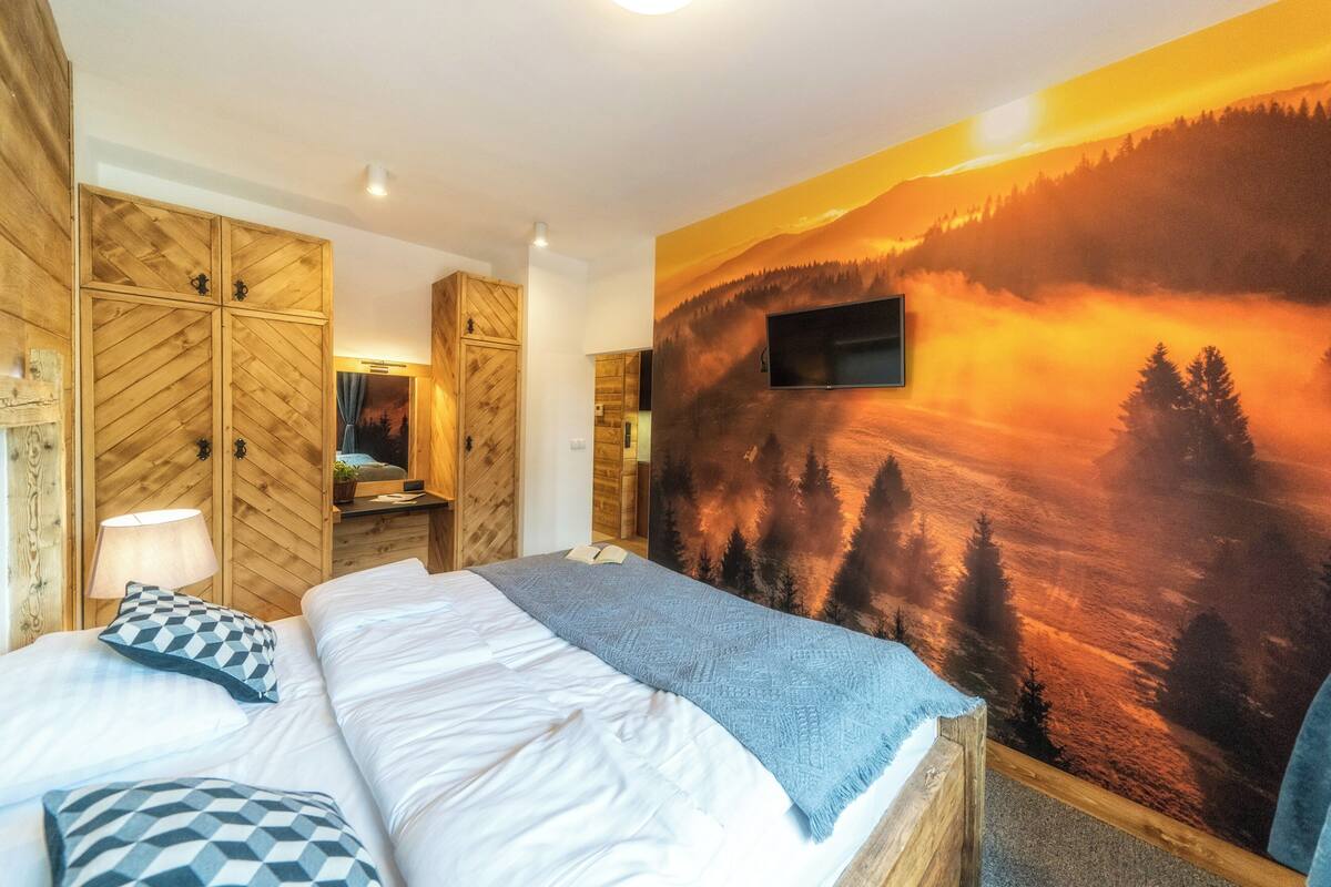 A bedroom with a bed, wooden wardrobe, a TV mounted on the wall, and a scenic sunset mural.