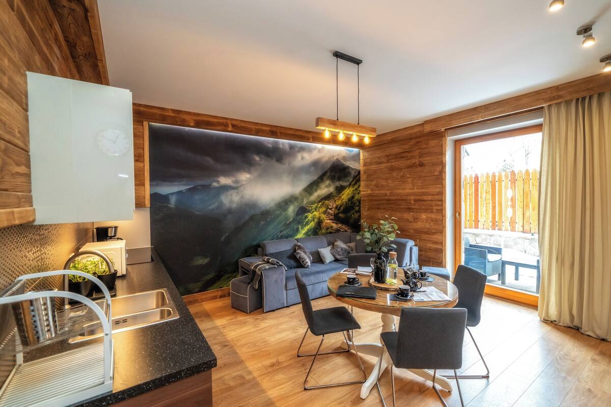 A modern living room with a large wall mural, a dining area with a glass table, and a kitchen with a sink and countertop.