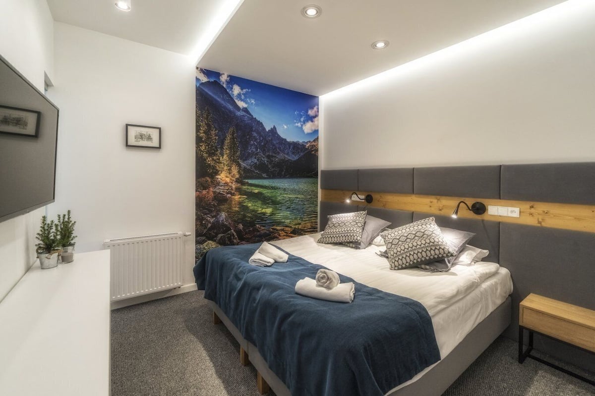 A modern bedroom with a large bed, a mounted TV, a radiator, and a scenic wall mural.