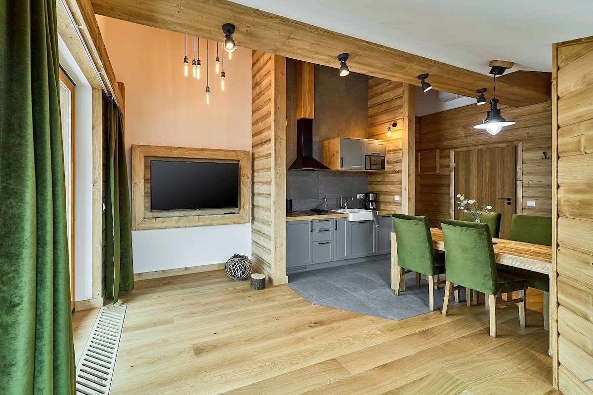 A cozy cabin-style room with a wooden interior, a dining area with green upholstered chairs, and a kitchen with modern appliances.