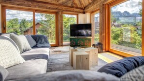 Una sala de estar acogedora con un televisor grande, muebles de madera y amplias ventanas con vistas a las montañas.