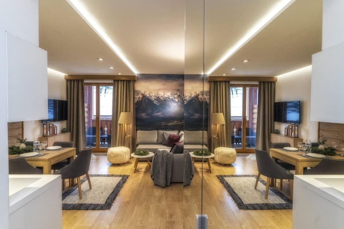 A modern hotel room with a large bed, a sofa, two armchairs, a dining table, and a TV.