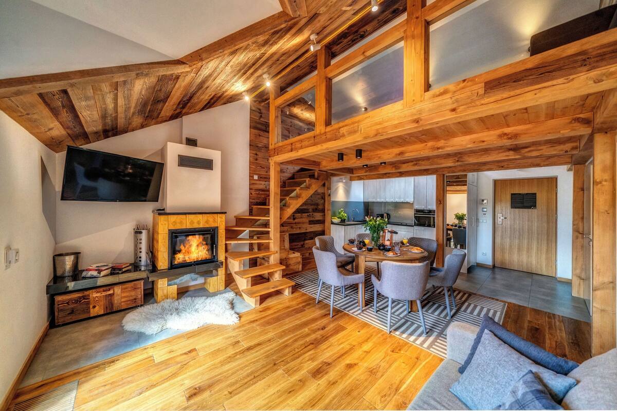 A cozy cabin interior with a fireplace, wooden staircase, dining area, and kitchen.