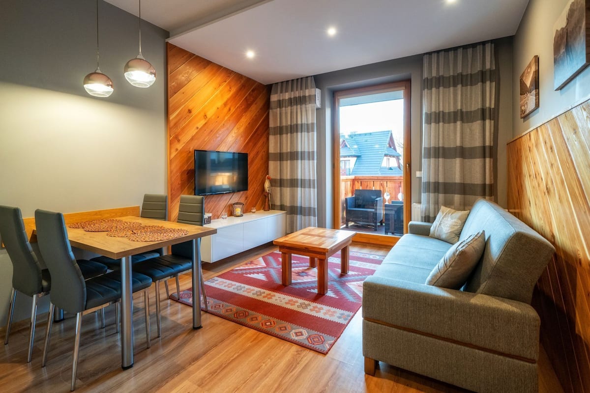 A modern living room with a wooden accent wall, a flat-screen TV, a sofa, and a dining area with chairs and a table.