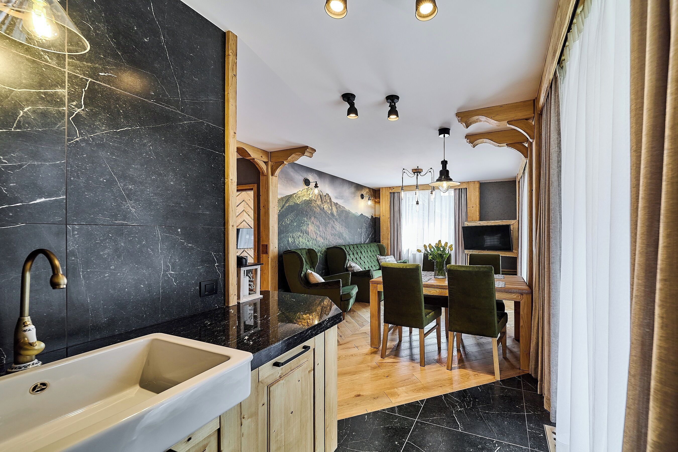Una cocina moderna con encimera de mármol, armarios de madera y un lavamanos. Hay una zona de comedor con mesa y sillas de madera, y un sofá verde.