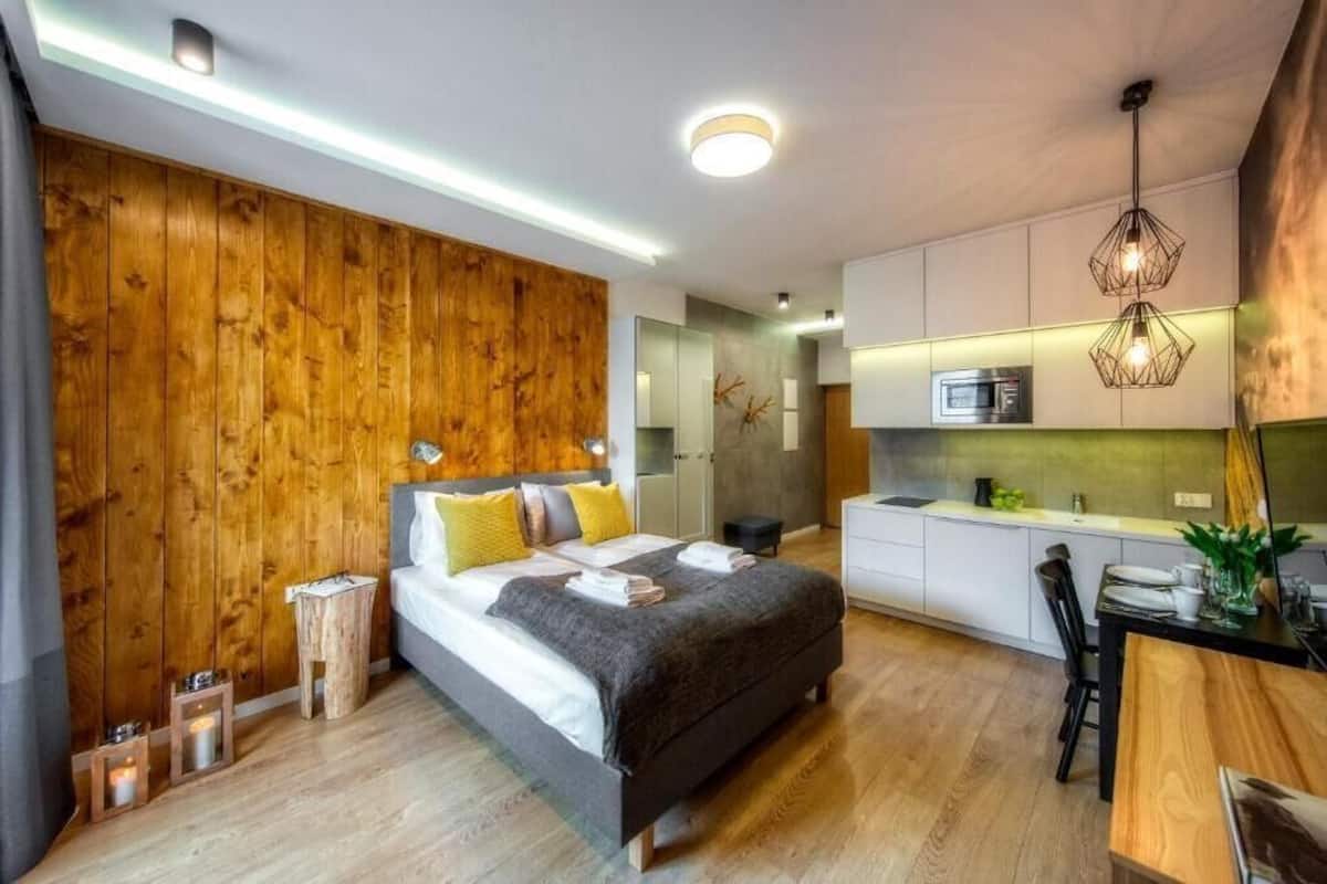 A modern bedroom with a wooden accent wall, a large bed with pillows, bedside tables, and a small bedside lamp. Adjacent is a compact kitchen with a microwave, a sink, and a dining area with a table and chairs.