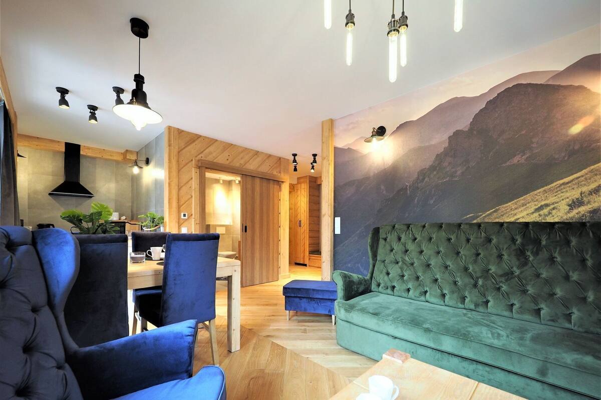 A modern living room with a green sofa, a blue armchair, and a wooden coffee table. There is a dining area with a table and chairs, and a kitchen with wooden cabinets and a black range hood. A large mountain mural is on the wall.