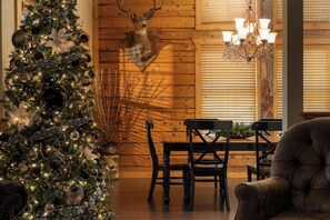 Een kerstboom met versieringen en lichtjes, een gewei aan de muur, houten wanden, een kroonluchter en een eettafel met stoelen.