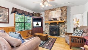 A cozy living room with a stone fireplace, wooden furniture, and a large window with blinds.