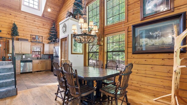 A wooden interior with a dining table surrounded by chairs, a chandelier, and a Christmas tree.
