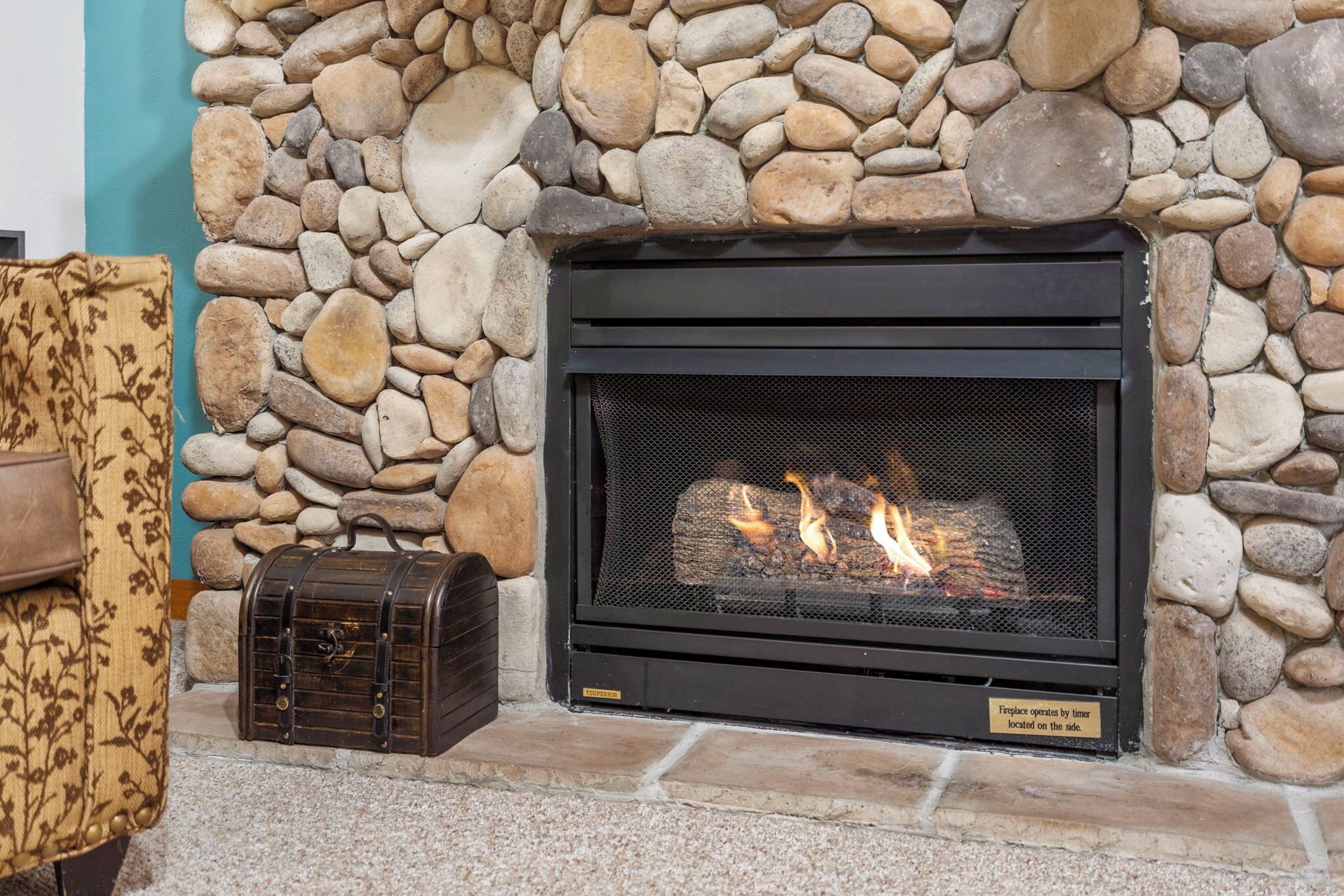 A stone fireplace with a black metal screen, a log fire, and a vintage suitcase on the left.