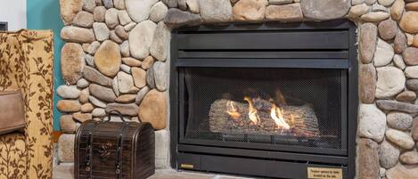 A stone fireplace with a black metal screen, a log fire, and a vintage suitcase on the left.
