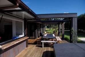 Eine überdachte Terrasse im Freien mit Holzbänken und einem Tisch, einer Pergola darüber und Blick auf eine Rasenfläche.