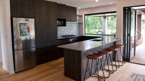 Eine moderne Küche mit dunklen Holzschränken, einem Edelstahlkühlschrank, einer großen Kochinsel mit Barhocker und Blick ins Grüne durch die Glastür.