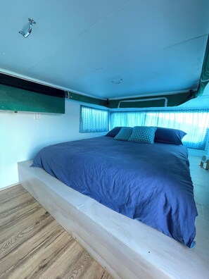 Ein Bett mit Holzbettgestell und blauer Matratze in einem Raum mit schrägem Dach und einem Fenster mit gemusterten Vorhängen.