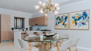 Una cocina moderna con armarios de madera, una mesa de comedor de vidrio y sillas blancas. Hay dos cuadros abstractos en la pared.