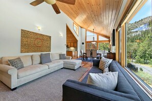 A spacious living area with a large sectional sofa, a smaller sofa, and a dark armchair. There is a wooden ceiling fan, a large window offering a view of a mountainous landscape, and a kitchen area visible through an open door.