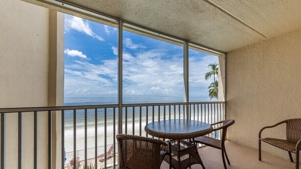 Een balkon met een ronde tafel en rieten stoelen, met uitzicht op een strand met palmbomen en de oceaan.