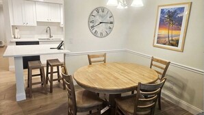 Een moderne keuken met witte kasten, een houten eettafel met vier stoelen, een klok en een lijst met een foto aan de muur.