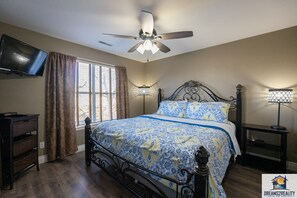 A bedroom with a bed, a ceiling fan, a TV mounted on the wall, and a window with curtains.