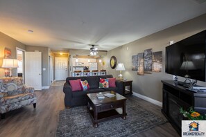 A living room with a dark sofa, a coffee table, and a TV. There is a kitchen in the background with a microwave and refrigerator. - Easy Access 2BR Branson Condo Near the Strip + Pool | Club C-6 (Branson)