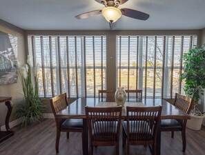 A dining area with a table and chairs, a ceiling fan, and large windows with blinds.