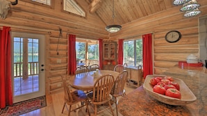 A wooden cabin interior with a dining table, chairs, a clock, and a bowl of apples. - Scenic Riverfront Retreat w/ Hot Tub & Kayaks! (Thompson Falls)