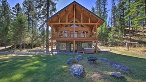 A two-story log cabin with a balcony, surrounded by a grassy yard and large rocks. - Scenic Riverfront Retreat w/ Hot Tub & Kayaks! (Thompson Falls)