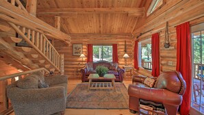 A log cabin interior with a wooden staircase, two sofas, a coffee table, and large windows with red curtains. - Scenic Riverfront Retreat w/ Hot Tub & Kayaks! (Thompson Falls)