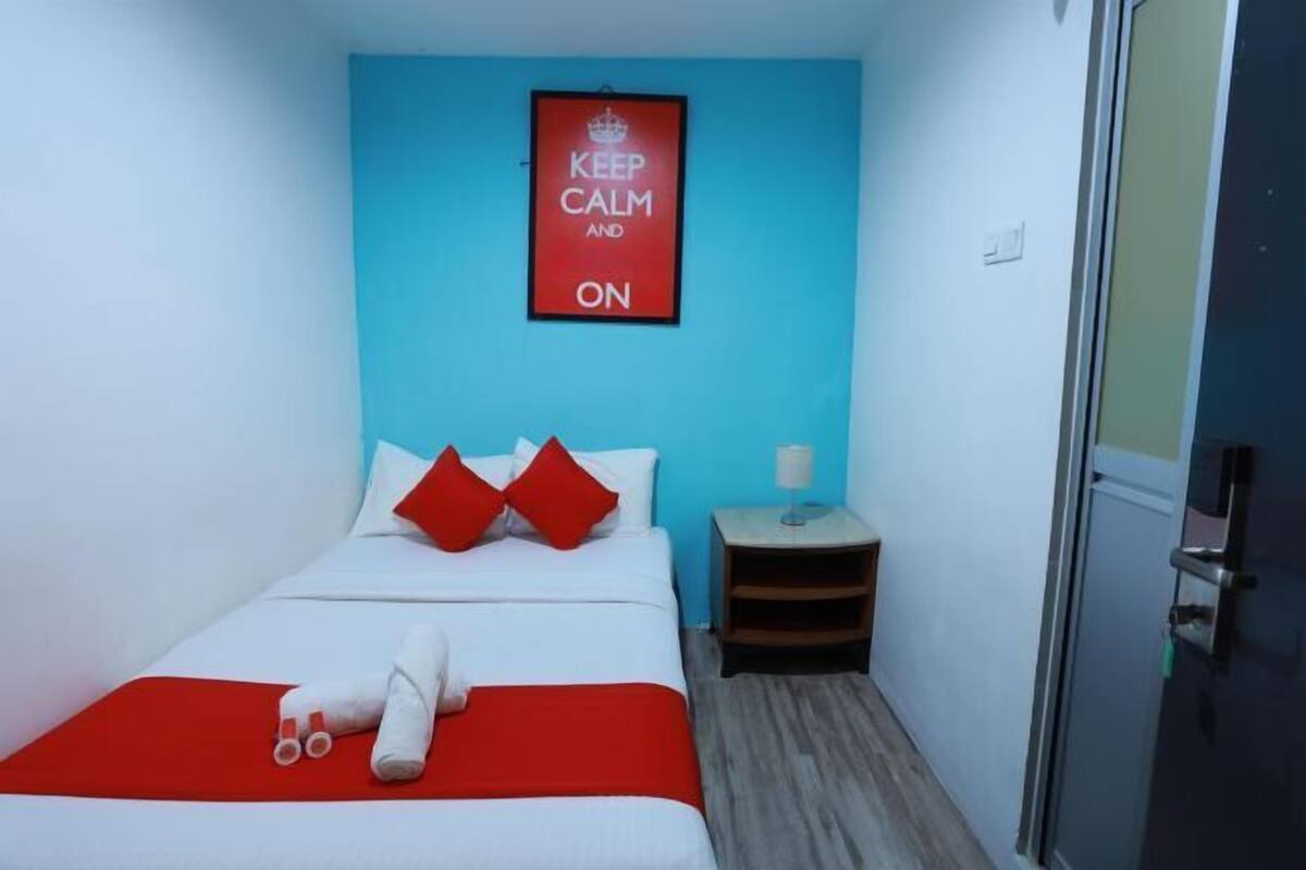 Ein Hotelzimmer mit einem Bett, rot-weiß gestreifter Bettwäsche, einem roten Kopfkissen, einem kleinen Nachttisch mit Lampe und einem Plakat mit der Aufschrift 'KEEP CALM AND ON' an der Wand.
