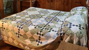 A wooden-paneled bedroom with a checkered quilt, a window with a view of trees, and a ceiling fan.