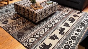 A cozy living room with a wooden interior, a black leather sofa, a patterned ottoman, and a large rug with animal prints.