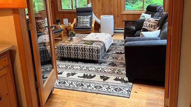 A cozy cabin interior with a wooden ceiling, a ceiling fan, a large window, a sofa, a chair, and a small table with a plant.