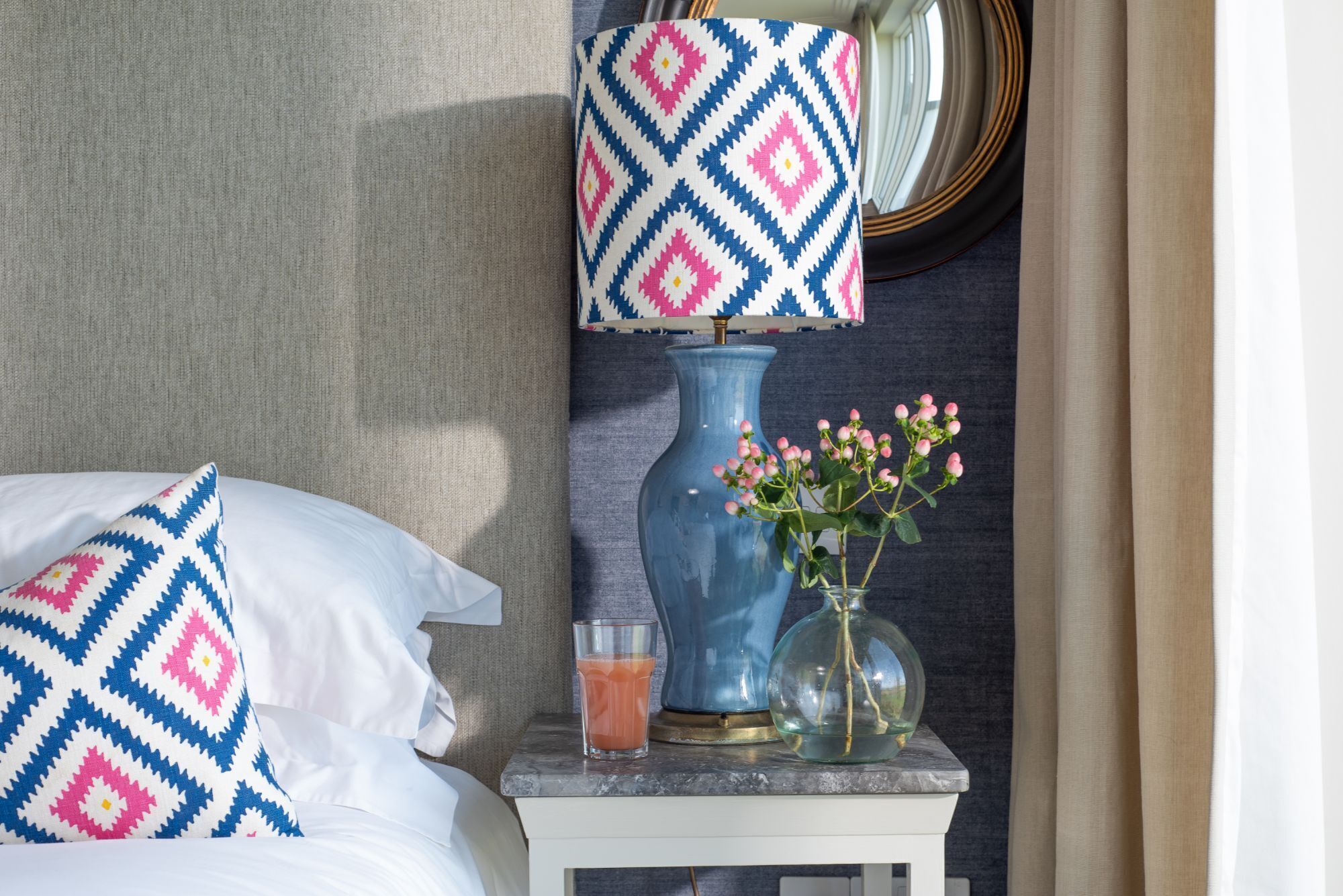 A neatly made bed with a patterned pillow, a bedside table with a vase of flowers, a glass of orange juice, and a blue vase.