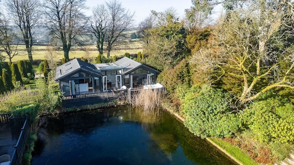 Une maison dotée d’une extension en verre surplombant une rivière, entourée d’arbres et de verdure.