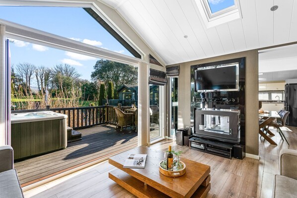 A spacious living area with a large TV, wooden flooring, and a view of the garden. - Berrynarbor Lodge, Kentisbury Grange (Barnstaple)