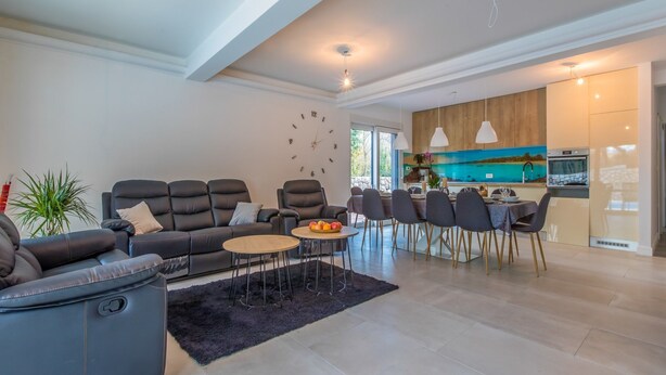 A modern living room with a sofa, a coffee table, and a dining area with chairs and a table. There is a kitchen visible in the background with a large window. - Villa Adry (Bribir)
