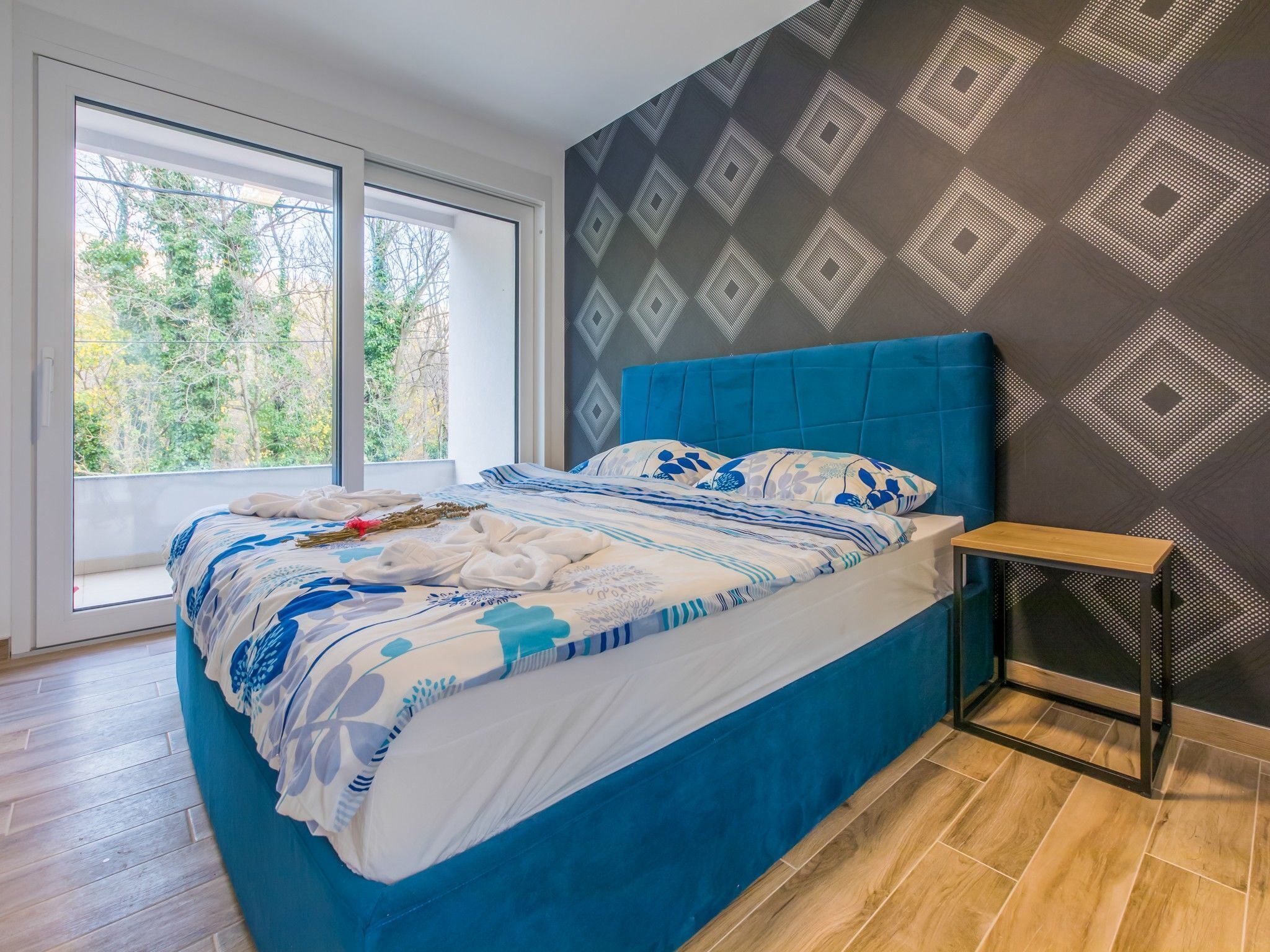 Un dormitorio moderno con una cama grande, una mesita de noche pequeña y una ventana con vistas a árboles.