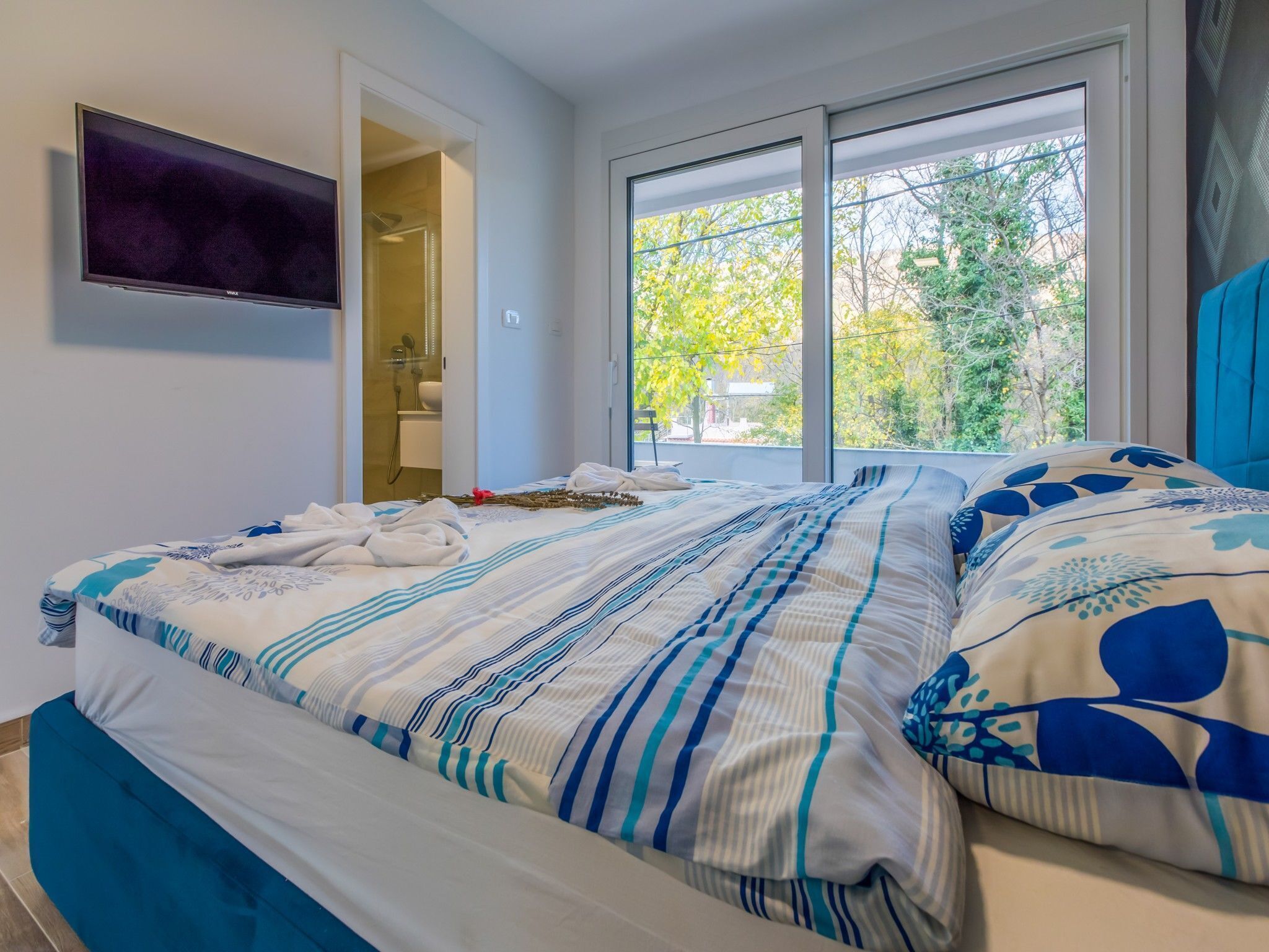 A hotel room with a bed, a TV mounted on the wall, a bathroom with a shower, and a window with a view of trees.