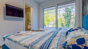 A hotel room with a bed, a TV mounted on the wall, a bathroom with a shower, and a window with a view of trees. - Villa Adry (Bribir)