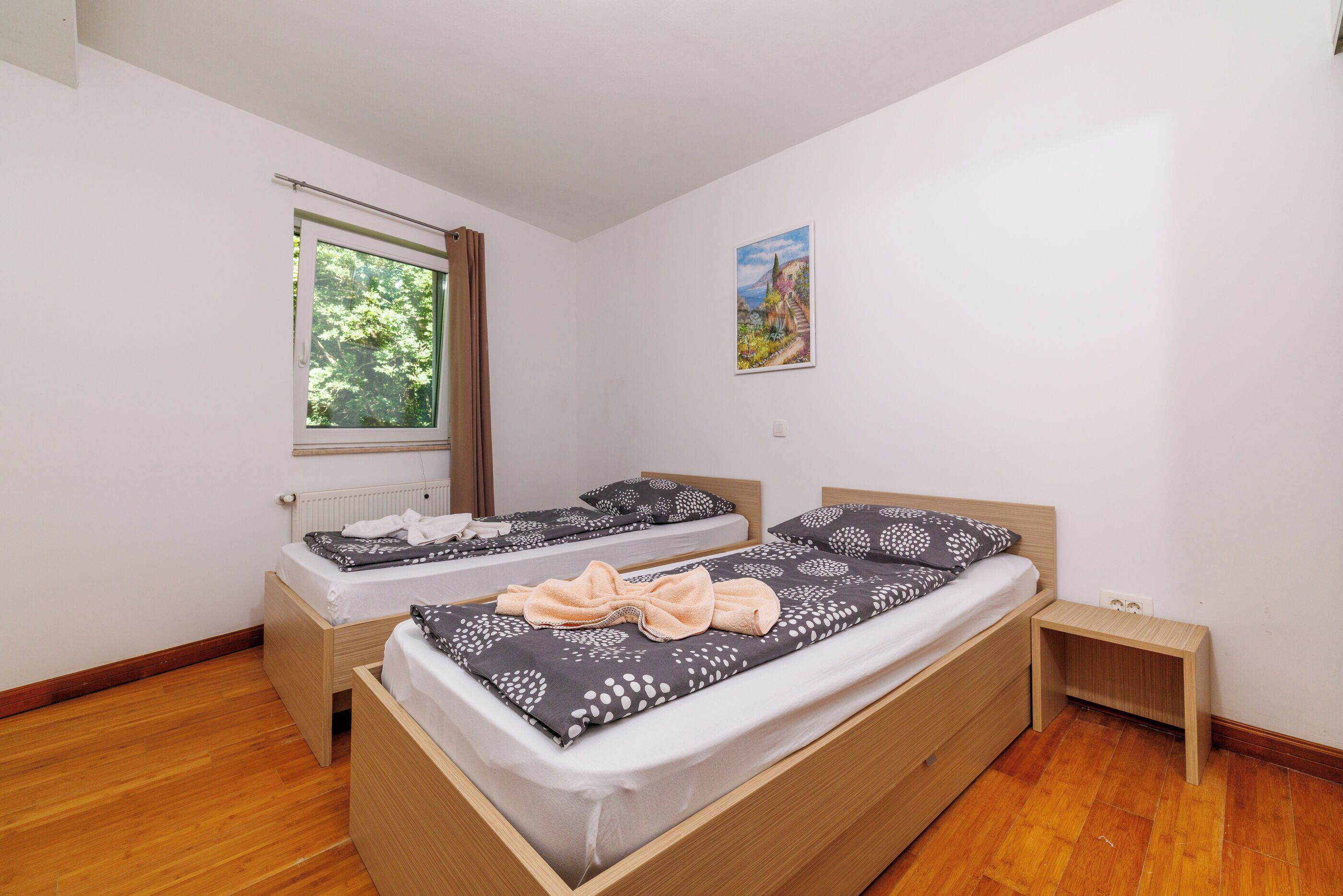 A bedroom with two beds, a window, a radiator, and a painting on the wall.