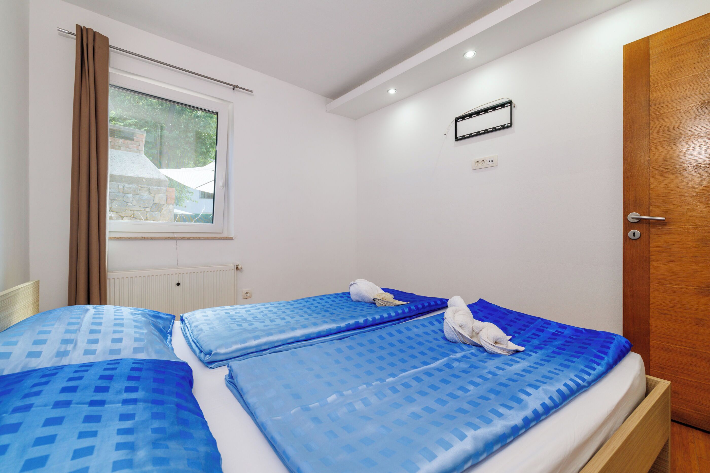 A hotel room with two beds, a window with curtains, a radiator, a wall-mounted TV, and a wooden door.