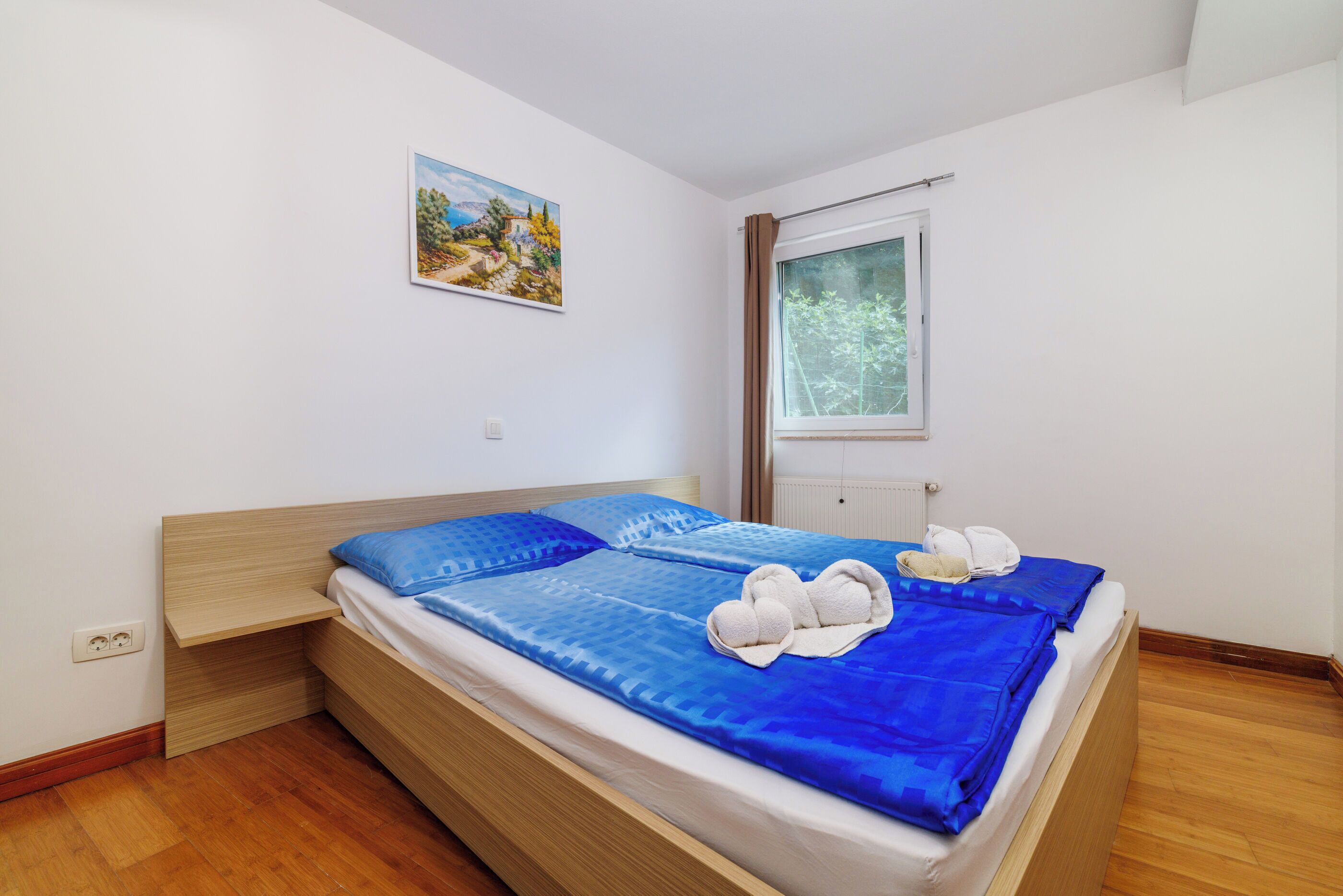 Un lit bien fait, avec une literie bleue, une tête de lit en bois, un tableau accroché au mur et une fenêtre avec des rideaux.