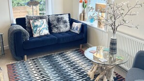 Un salon moderne avec un canapé bleu foncé, des coussins à motifs, une table basse en verre et une vue sur la mer grâce à de grandes fenêtres.